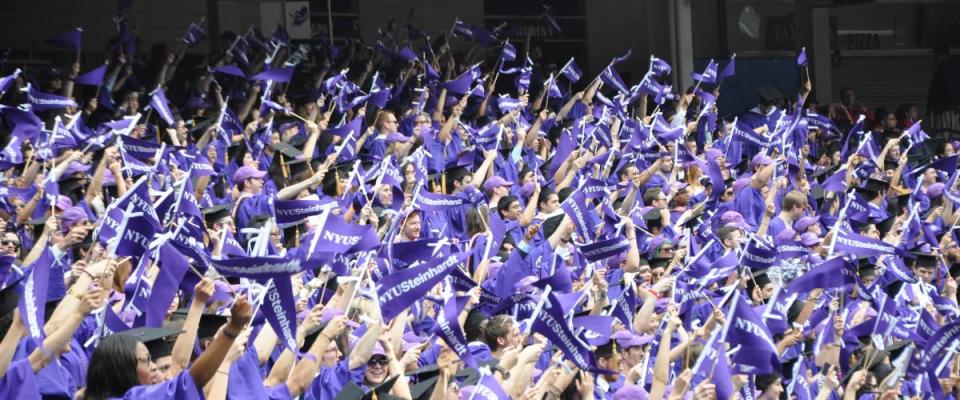 NYU graduation, students in purple caps and gowns waving NYU flag