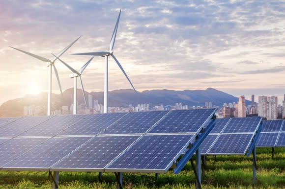 Wind towers and solar panels with a city in the background.