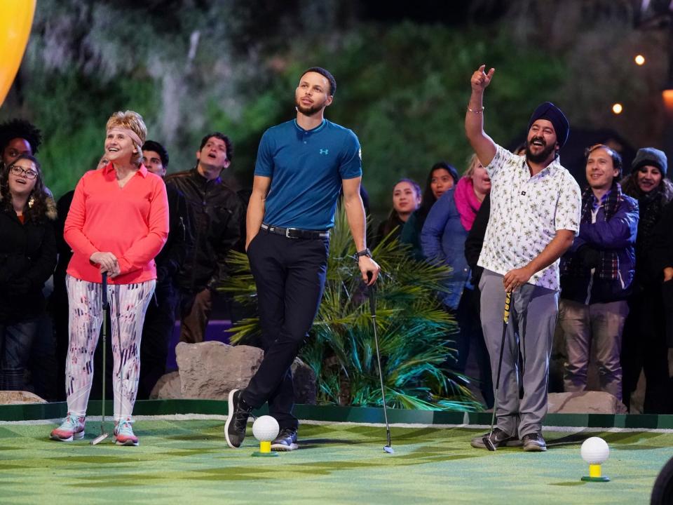 Stephen Curry leans on a golf clup and looks on besides contestants and a crowd on the set of "Holey Moley."