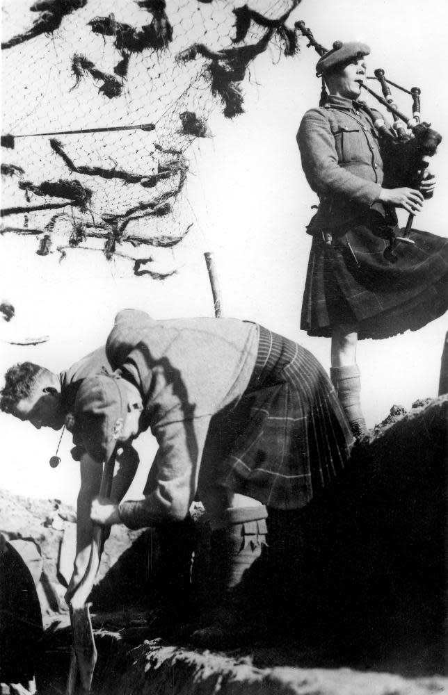 A detachment of the Highland Regiment dig trenches in France during the second world war, accompanied by a piper.
