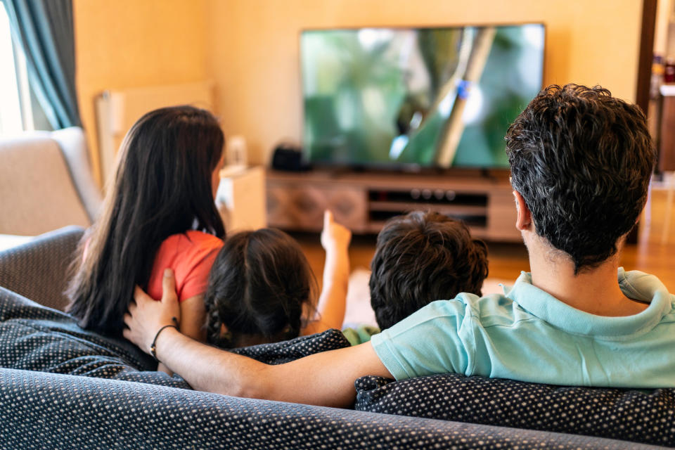 Two kids sit with their parents and watch TV on the couch