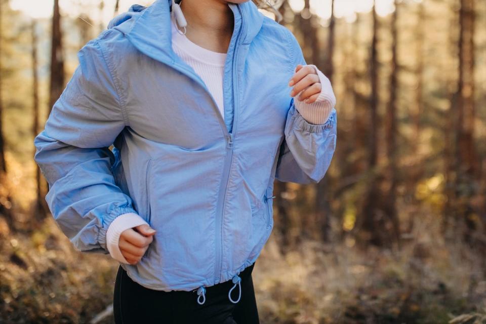 A woman runs along a wooded trail while wearing a powder blue lightweight jacket.