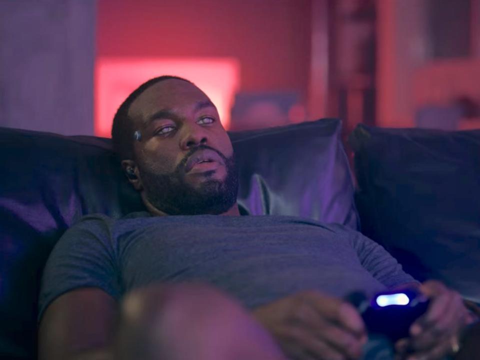 yahya abdul-mateen II as karl in black mirror, laying back on a couch with a vacant expression on his face. he's holding a video game controller and his eyes are glowing