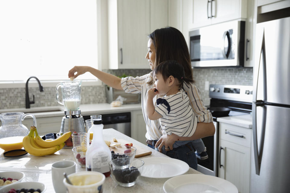 Mother holding baby daughter, blending smoothie in kitchen