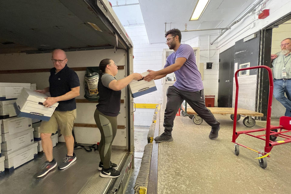 Three people on a loading dock handle white cardboard boxes in the back of a truck as someone leans against a doorway watching them..