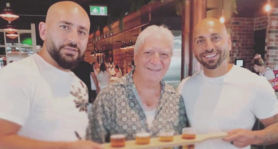 The father and sons stand holding beers while smiling at the camera. 