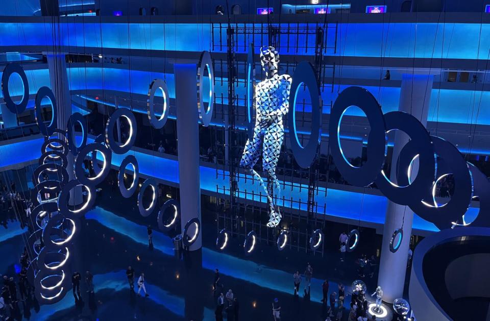 A holographic image of a human figure is seen between rows of ring lights in a space-age atrium.