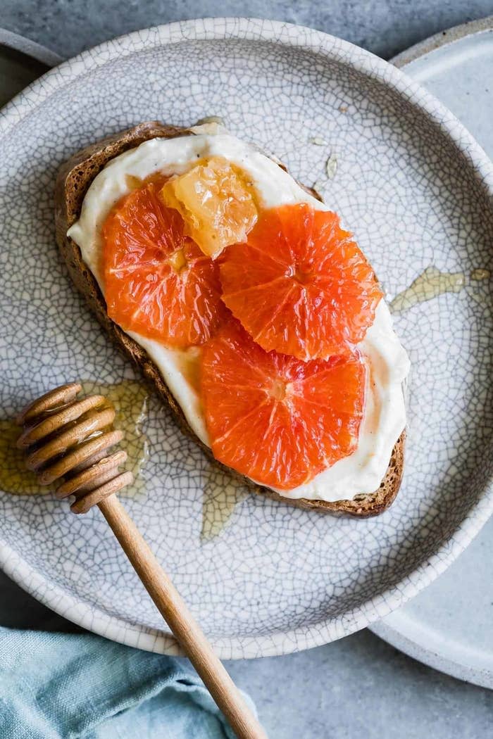 A slice of bread with cream cheese and citrus fruit on a plate, with a honey dipper alongside