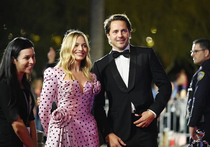 Margot Robbie in a stylish polka dot dress with Tom Ackerley in a black tuxedo on the red carpet, smiling for photos. Another unidentified smiling woman stands beside them