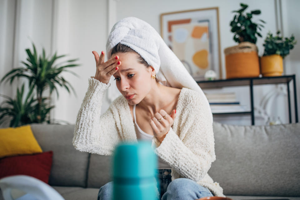 One woman, beautiful young woman sitting on sofa in living room and woman applying moisturizer on her face, she has a towel on her head.