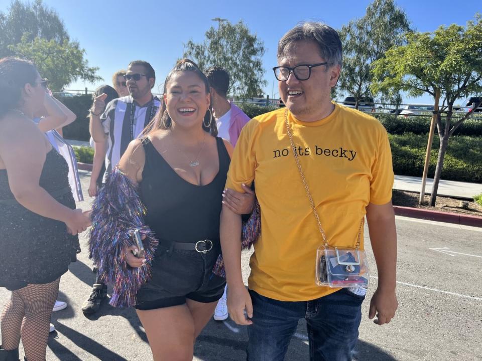 A woman in black shorts and top and a man in a T-shirt that says "no its becky" stand outside a concert venue.