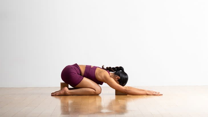 A woman practices Balasana (Child's Pose) with blocks under her head and hips. She is wearing a burgundy athletic top and shorts. Kneeling on a light wood floor against a white background.