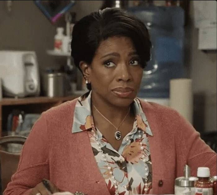 I don't know who is in the image. A person in a patterned blouse and cardigan sits at a table with a skeptical expression. Various household objects are in the background