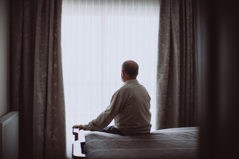 An old senior man facing a window while sitting on a bed.