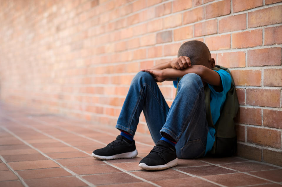 A young boy in a blue shirt and jeans sits on the ground against a brick wall, with his head resting on his crossed arms