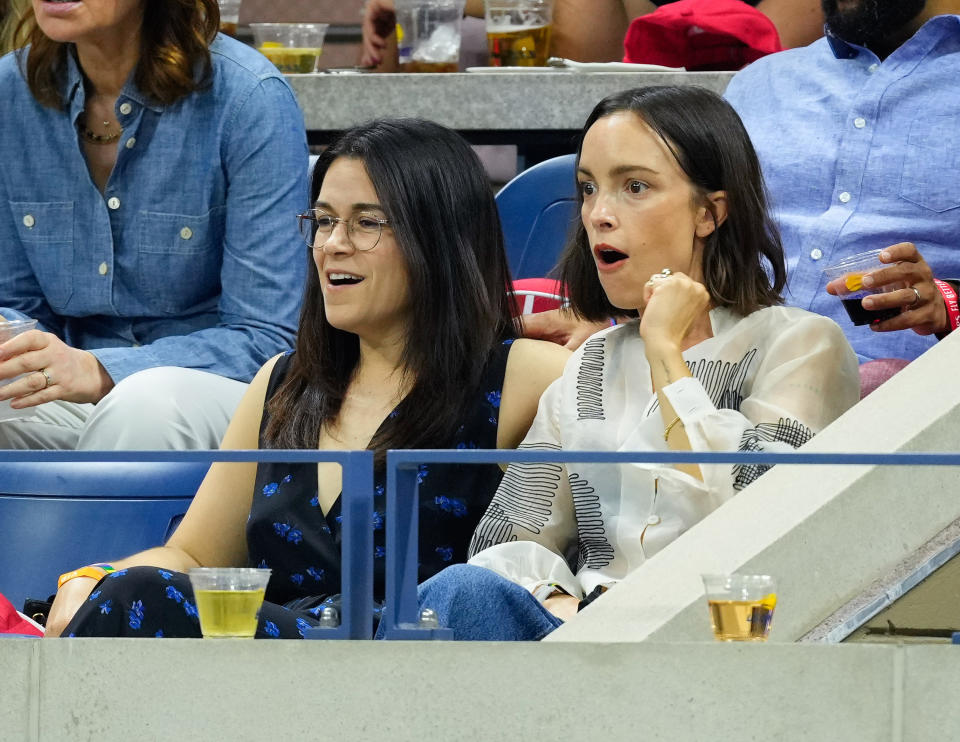 Abbi Jacobson and Jodi Balfour sit together at a sports event. Abbi wears a patterned outfit while Jodi wears a shirt with black detailing. Both are watching intently