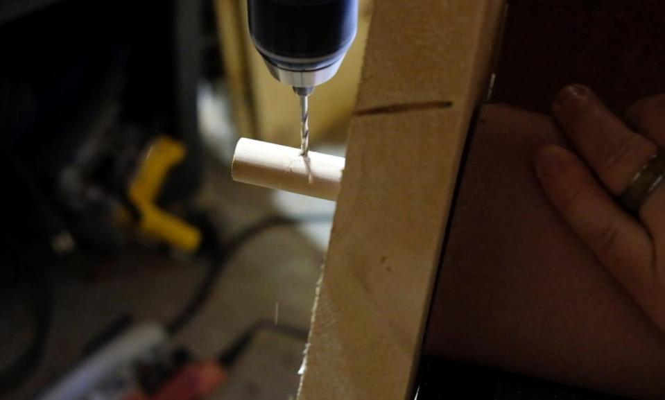 A man drilling a small hole in the end of a dowel in a wall-mounted paper roll holder, to install a cotter pin so the dowel will stay in place.