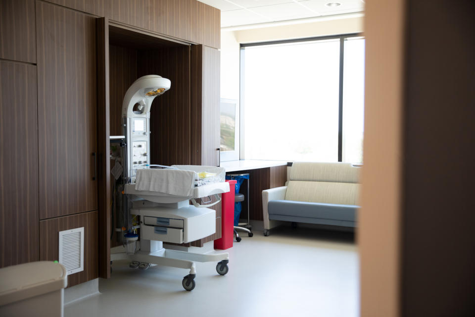 Hospital room with a baby bassinet and medical equipment, a couch near a large window, and wooden cabinets. Room is empty of people