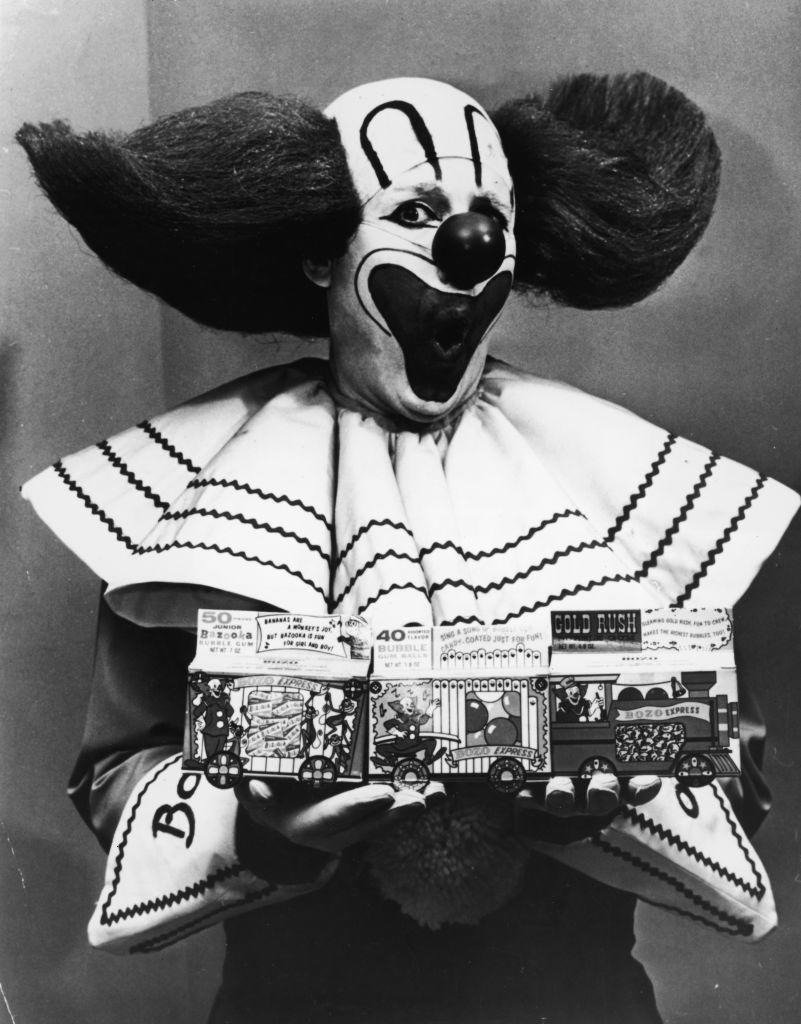 A vintage photo of Bozo the Clown holding two toy boxes labeled "50 Tricks" and "40 Tricks" with "Gold Rush" written on one. Bozo is in his iconic clown costume