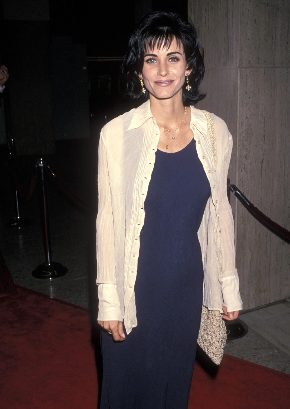 Courteney Cox on the red carpet wearing a long-sleeve shirt over a long dress, accessorized with earrings and a textured clutch
