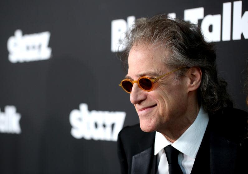 Richard Lewis wears yellow-rimmed sunglasses and smiles in a suit at a premiere