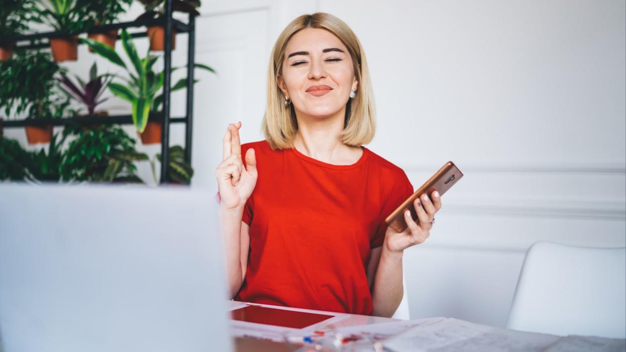  young woman with dyed blonde hair and a bright red shirt sits in front of a laptop; her eyes are closed and her fingers are crossed, as if she's making a wish. In one hand, she holds a cell phone. 