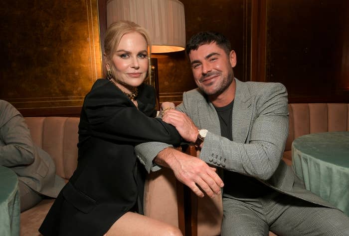 Nicole Kidman, in a chic black ensemble with gold jewelry, and Zac Efron, in a grey suit, are sitting together in an elegant setting, smiling at the camera