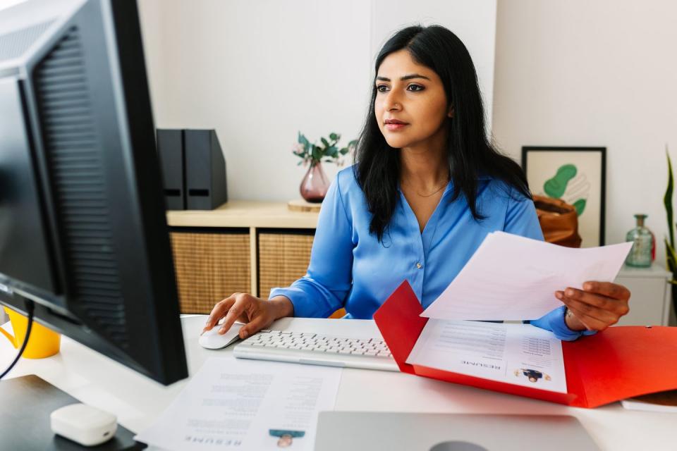 young indian woman holding a cv and looking at a computer screen