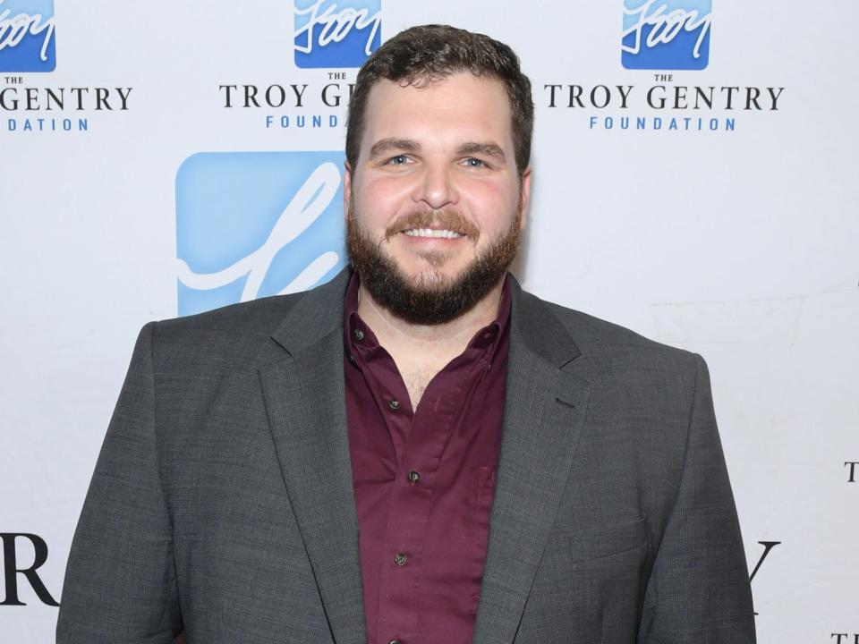 Jake Hoot wearing a gray blazer and red shirt in front of a white and blue background