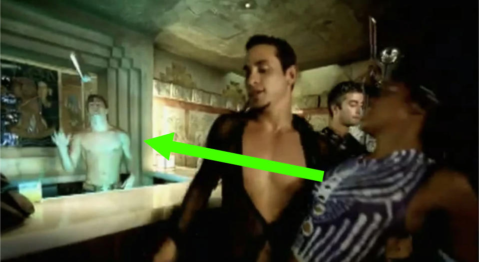 A still from a music video, set in a bar, there's a shirtless bartender throwing a cocktail shaker and an arrow pointing at him