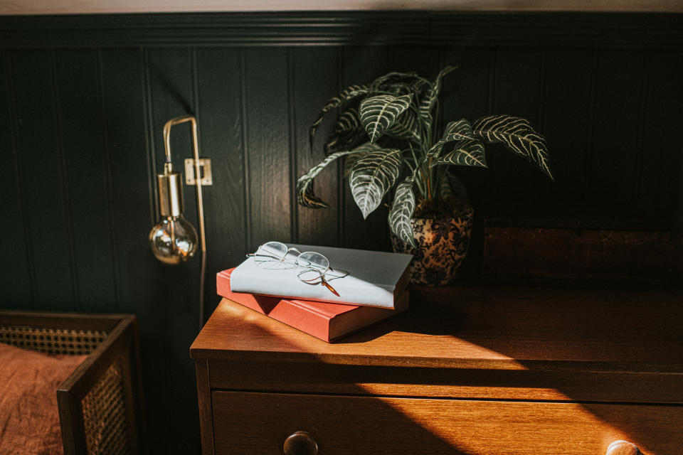 A red and a white book placed on a mid-century style bedside table, bathed in sunlight. A simple pair of reading spectacles rest onto of the white book, creating a shadow. An exposed bulb wall mounted light is visible, and a house plant is obscured in shadow.