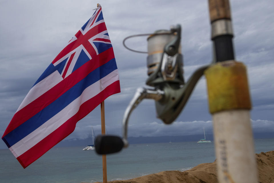 A fishing pole and the Hawaiian state flag are posted on the popular tourist beach of Kaanapali on Friday, Dec. 1, 2023, in Lahaina, Hawaii. Lahaina Strong has set up a "Fish-in" to protest living accommodations for those displaced by the Aug. 8, 2023 wildfire, the deadliest U.S. wildfire in more than a century. More than four months after the fire, tensions are growing between those who want to welcome tourists back to provide jobs and those who feel the town isn't ready for a return to tourism." (AP Photo/Ty O'Neil)