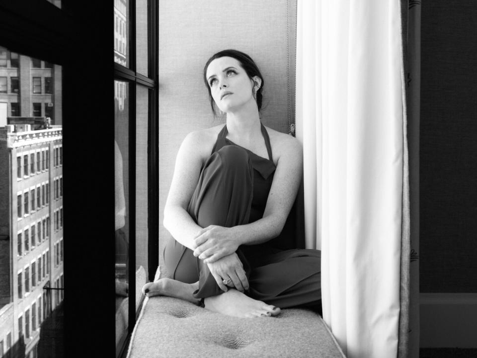 A woman sits on a bench against a window and looks up.