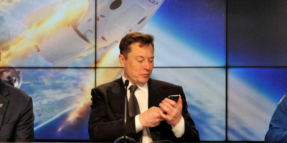 Elon Musk looks at his phone with a display of a space capsule entering orbit behind him.