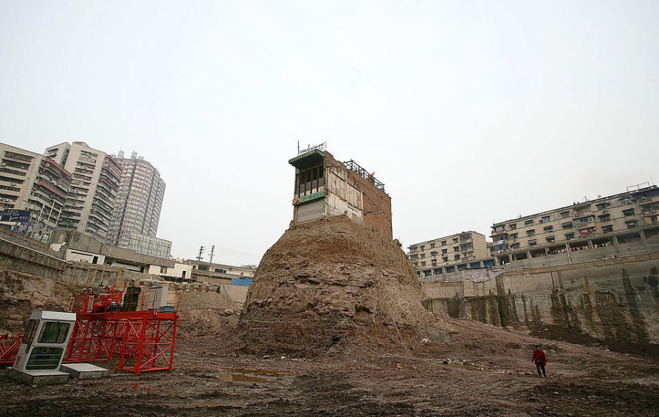 A lone building stands atop an isolated dirt mound amidst a construction site surrounded by apartment buildings