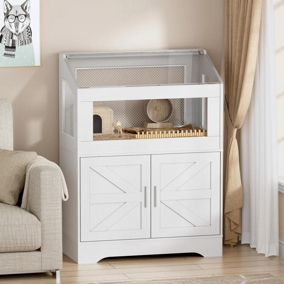 Stylish white wooden hamster cage with a large viewing window, exercise wheel, and tunnel system, placed in a cozy, living room setting