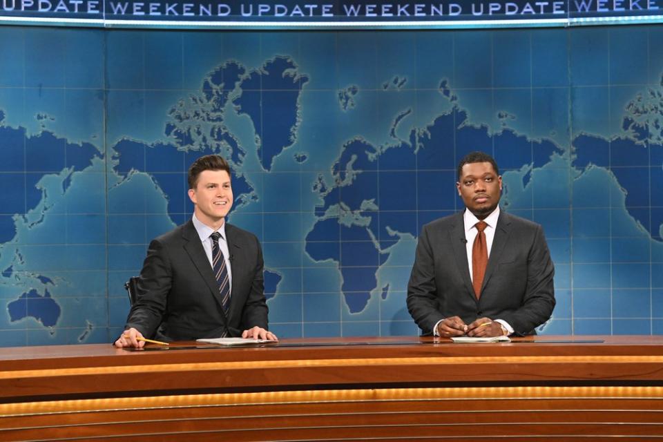 Two men in gray suits at a news anchor desk, in front of a map of the world