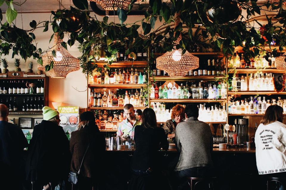 large bar with bottles on gold, glass shelves, bartenders making drinks, and people sitting at bar talking