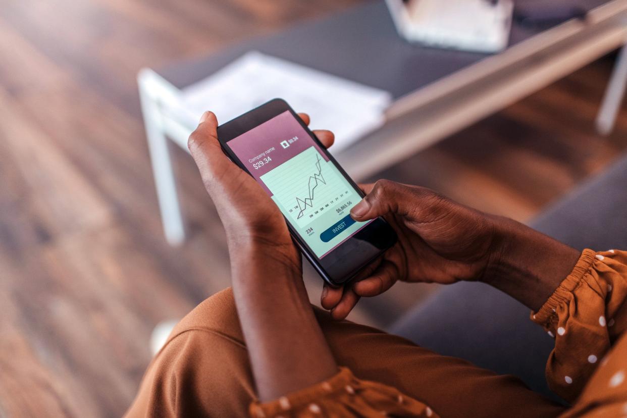 Closeup of woman's hands holding smartphone looking at stocks while sitting on couch, blurred table and floor in the background