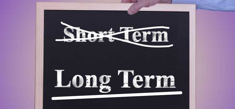 Hand holding chalkboard with words long term and short term with long term underlined and short term crossed through.