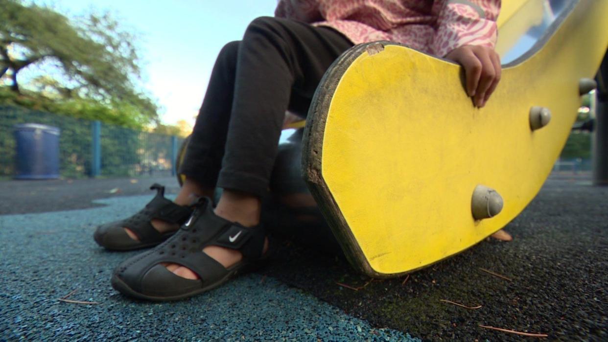 Aiyla, a child in black shoes, black leggings and a pink coat, whose face is not visible, sits at the bottom of a yellow slide with their feet touching the blue and black floor