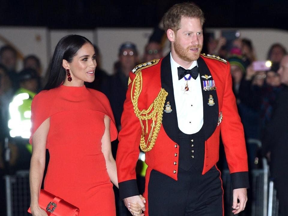 Meghan in a red cape sleeve dress and Harry in a red military jacket with badges, ropes, and a bow tie.