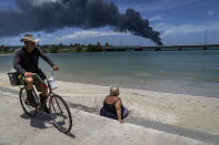 A man rides his bicycle while a sunbather looks out at as column of smoke rises from the Matanzas supertanker base in Matanzas, Cuba, on Sunday, Aug. 7, 2022. Cuban authorities say lightning struck a crude oil storage tank at the base, sparking a fire that sparked four explosions that injured more than 121 people, with one person dead and 17 missing. (AP Photo/Ramon Espinosa)