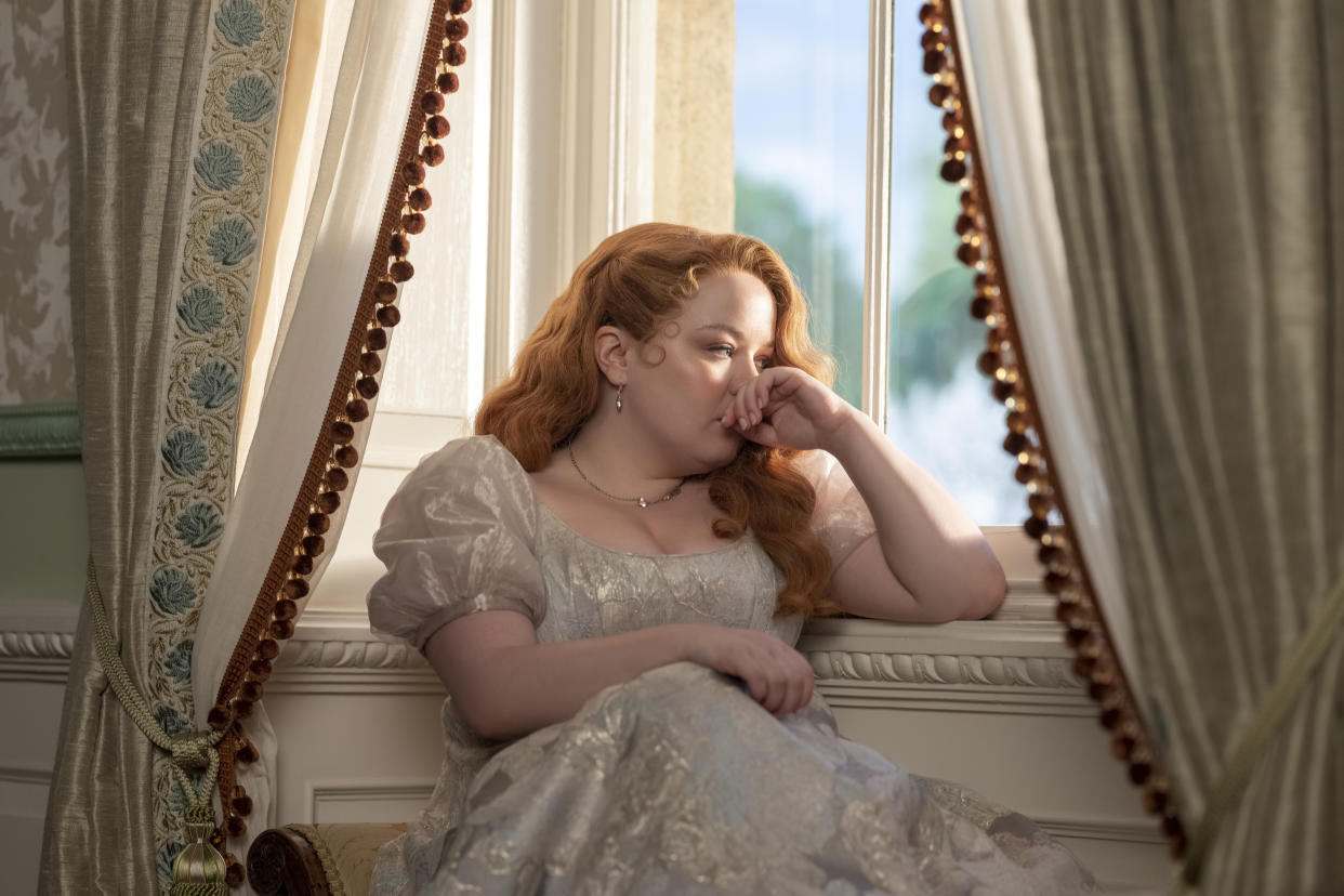 Nicola Coughlan, as Bridgerton character Penelope Featherington, sits by a window with her hand to her mouth.