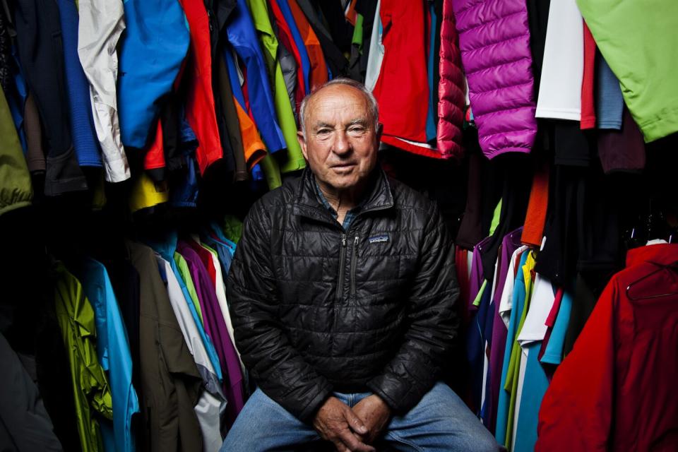 An older man in a dark jacket sits in front of other apparel.