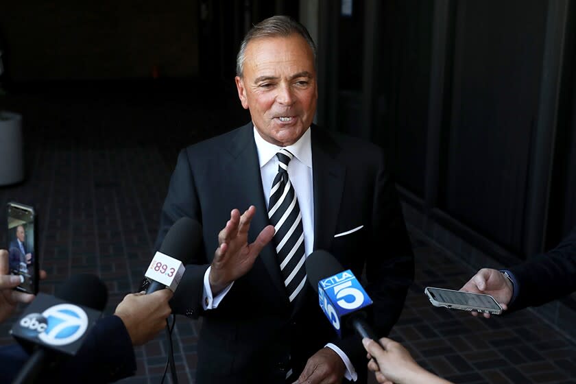 A man in a suit and tie faces a line of hands holding microphones with TV station logos on them.