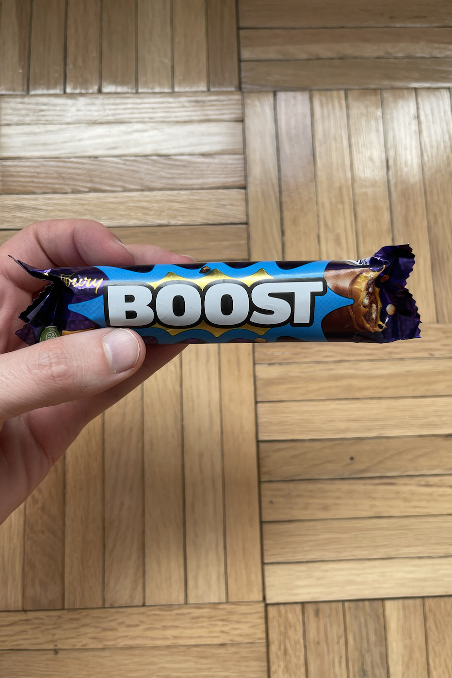 Hand holding a partially unwrapped Boost chocolate bar over a wooden floor