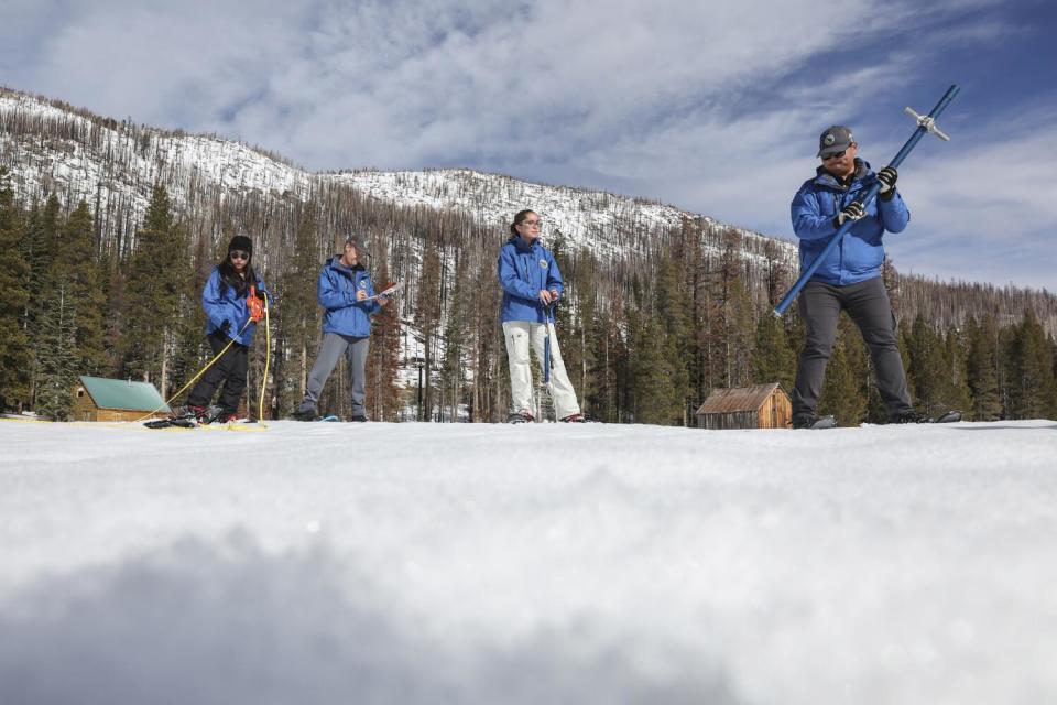 People in blue parkas stand in a field of snow holding a tube used to measure snow depth.