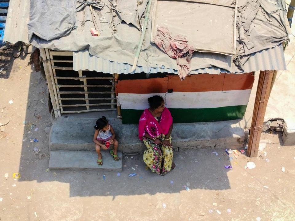 A woman and young girl sit in shade outside a small building with slats in the doors and a precarious looking roof.