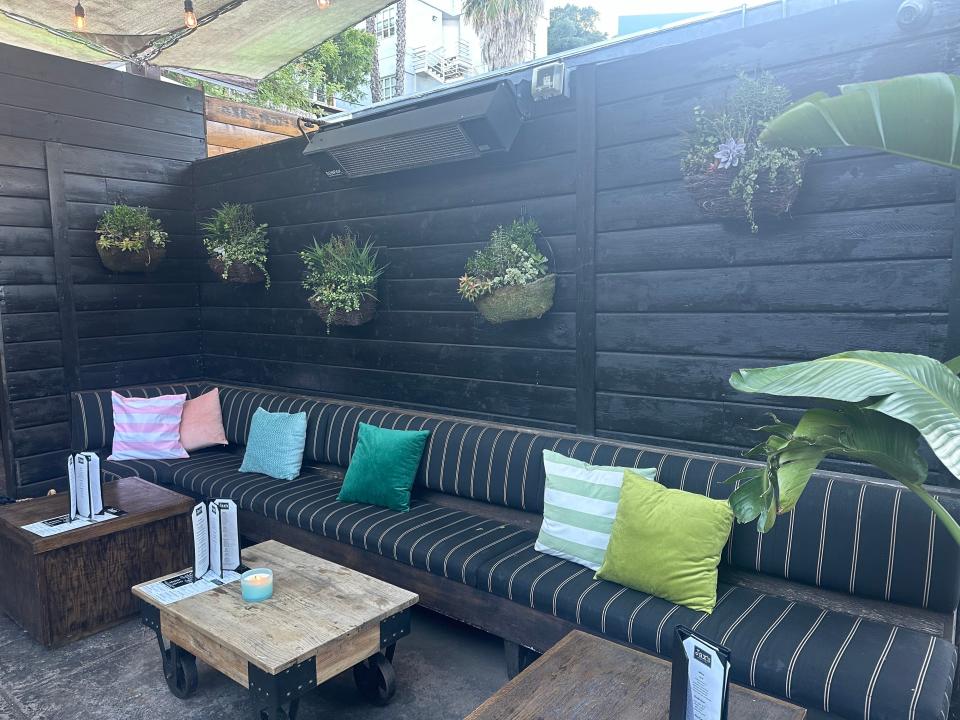 Outdoor dining area with a long bench, pillows, and wooden tables, ready for patrons